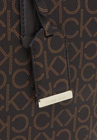 Borsa a tracolla marrone con texture e motivi a logo ripetuti. Presenta un'etichetta dorata con la scritta "Calvin Klein" e un accento in pelle marrone.