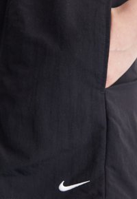 Tissu noir avec une texture lisse et une poche latérale. Présente un petit logo Nike blanc près de l'ourlet inférieur. Design minimaliste.