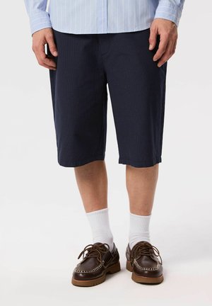 Mężczyzna w granatowych teksturowanych szortach, białych krótkich skarpetach, brązowych skórzanych mokasynach oraz jasnoniebieskiej koszuli w paski z guzikami.