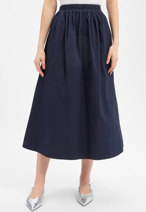 Kobieta w granatowej spódnicy midi z elastycznym pasem i srebrnych szpilkach z ostrym noskiem, stojąca na jednolitym białym tle.