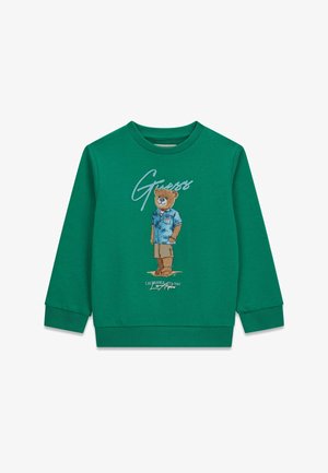 Felpa a maniche lunghe verde con grafica di un orso che indossa una maglietta blu, pantaloncini beige e sandali, con la scritta "Guess" sopra.