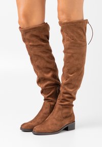 Bottes en suède marron au-dessus du genou avec un talon plat, un design décontracté et une fermeture zippée sur le côté. Texture lisse et détails de couture simples.