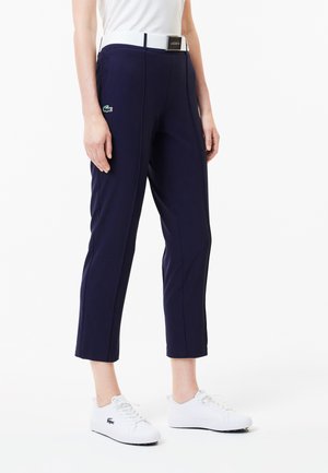 Navy cropped pants with a textured surface, featuring a white belt and green logo patch. Paired with white sneakers for a casual look.