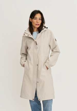 Femme portant un imperméable beige à capuche, longueur genou, avec des poches à l'avant et un jean bleu, debout devant un fond clair uni.