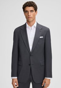 Mężczyzna w ciemnoszarym, strukturalnym garniturze z białą koszulą i białą poszetką, stojący na tle jednolitego, jasnoszarego tła.