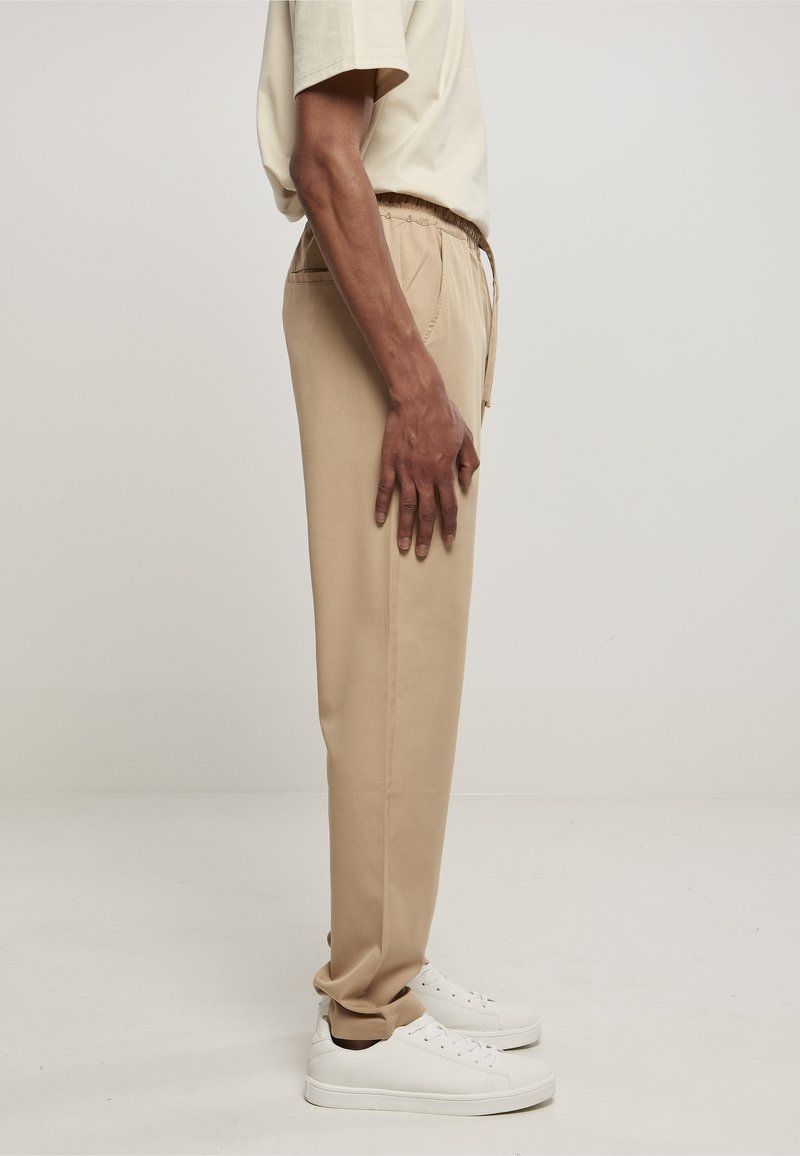 Personne en pantalon beige décontracté avec une taille élastique et des baskets blanches, debout de profil sur un fond uni.