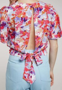 Blouse florale avec des manches bouffantes, présentant une découpe au dos et un détail à nouer à la taille. Les couleurs incluent du rose, du violet et de l'orange sur un fond clair.