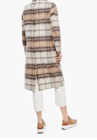 Abrigo largo de cuadros en crema, marrón y beige, hecho de tela texturizada. Presenta una abertura en la parte posterior y se combina con pantalones blancos cortos y zapatillas.