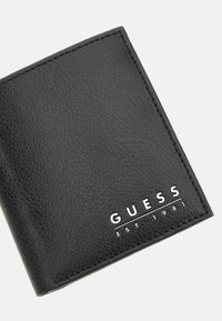 Portefeuille en cuir noir avec une finition texturée, arborant un logo blanc en relief "GUESS EST 1981" à l'avant et des bords nets et précis.