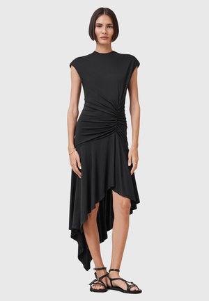 Robe asymétrique noire en tissu doux avec des manches courtes, dotée d'un détail frontal torsadé et d'un ourlet fluide. Associée à des sandales noires.