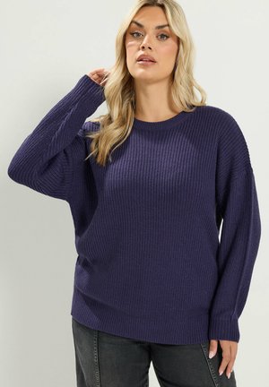 Donna con capelli biondi che indossa un maglione a maglia viola scuro e jeans scuri, in piedi davanti a uno sfondo chiaro e semplice.