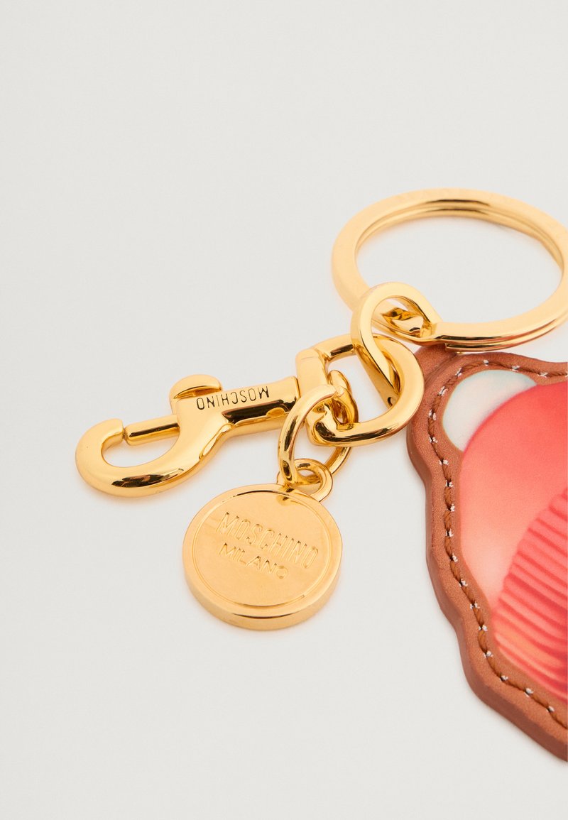 Guldtonad nyckelring med en hummerklämma och rund skiva märkt "MOSCHINO MILANO." Det finns en rosa och brun stiliserad björn design fäst vid den.