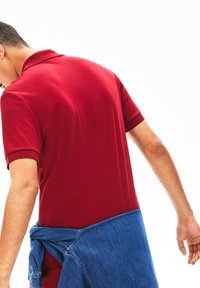 Czerwona bawełniana koszulka polo z krótkimi rękawami, o gładkiej fakturze. Niebieska dżinsowa kurtka zawiązana w talii. Prosty widok z tyłu.