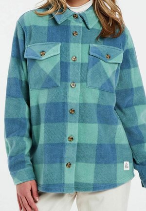 Femme portant une chemise en polaire à carreaux verts et bleus, boutonnée avec deux poches poitrine à boutons et une petite étiquette logo blanche à l'ourlet.