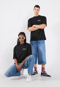 Two models wear matching black oversized t-shirts with text. The woman crouches in light blue jeans and white shoes; the man stands nearby in blue denim shorts and black sneakers.