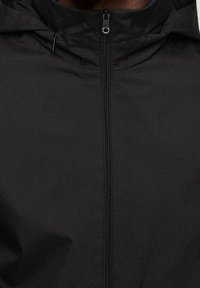 Veste noire avec fermeture éclair à l'avant, capuche et texture de tissu lisse. Design simple et minimaliste sans logos ni motifs visibles.