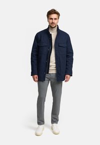 Marineblå jakke med to brystlommer, knaplukning foran, lagdelt over en lysebeige turtleneck, parret med grå bukser og hvide sneakers.