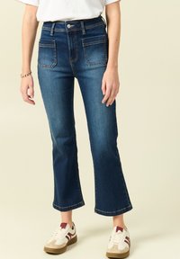 Ciemnoniebieskie spodnie dżinsowe z wysokim stanem i rozszerzanym dołem, z dwoma przednimi kieszeniami, jasnym przeszyciem i gładką fakturą denimu.