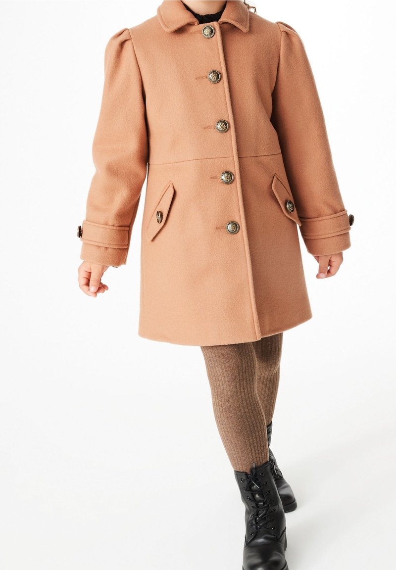 Abrigo de lana color durazno con cuello, cuatro botones en el frente, dos bolsillos laterales y mangas abullonadas, combinado con mallas marrones acanaladas y botas negras.