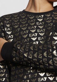 Felpa nera con motivo a logo EA7 dorato ripetuto. Il materiale sembra morbido con una finitura strutturata. Collo e polsini a costine visibili.