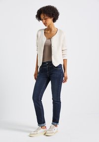 Cardigan beige clair texturé avec un col rond et une fermeture à boutons, associé à un jean bleu foncé et des baskets crème.