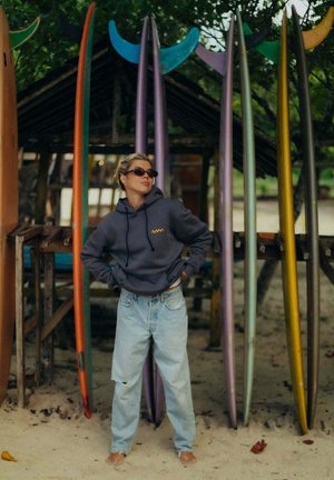 Osoba w okularach przeciwsłonecznych i kapturze stoi boso na piasku obok szeregu kolorowych desek surfingowych pod zacienioną drewnianą konstrukcją.