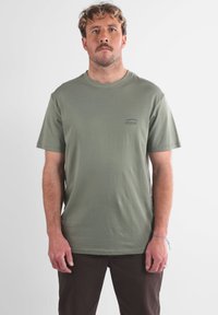 T-shirt en coton vert olive à manches courtes, col rond, avec un petit logo bleu sur la poitrine gauche. Porté avec un pantalon marron foncé.