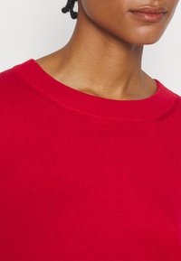 Gros plan d'une personne portant un pull en tricot rouge vif à col rond, montrant une partie du cou et des lèvres.