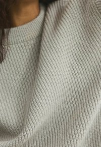 Pull gris clair en maille avec une texture côtelée et un large décolleté. Le tissu tombe doucement, révélant un motif subtil de la trame.