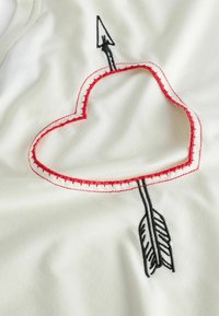 Witte stof met een rode geborduurde hartvorm met een zwarte pijl door het midden. Gladde textuur met gedetailleerde stitching.