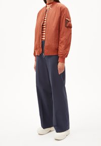 Giacca bomber color arancione ruggine con zip, polsini a costine e tasca cargo frontale, abbinata a pantaloni larghi blu navy e scarpe da ginnastica bianche.