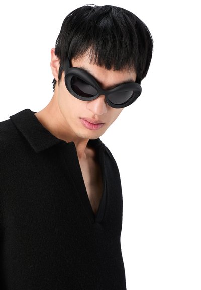Black oval sunglasses with a thick, matte frame; worn by a person in a black, textured shirt with a V-neck. Close-up perspective.