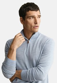 Mann mit kurzen braunen Haaren, der einen hellblauen Piqué-Pullover mit quartiertem Reißverschluss trägt, einen Kragen mit einer Hand hält und zur Seite schaut.