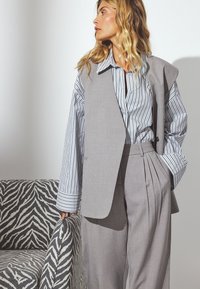 Gilet grigio oversize sopra una camicia a righe con bottoni, abbinato a pantaloni ampi con pieghe. Sullo sfondo si trova un divano a motivo zebra.