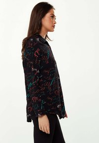 Black textured shirt featuring multicolored scribble designs in red, green, and white. Long sleeves with a collared neckline and side slits.