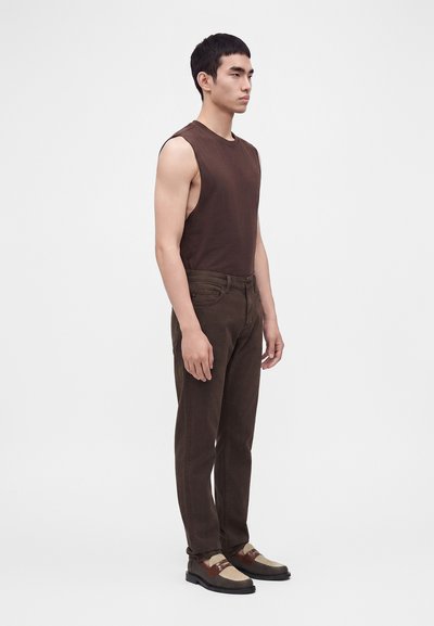 Jeune homme debout de profil portant un haut sans manches marron foncé, un pantalon marron foncé et des mocassins tissés bicolores.