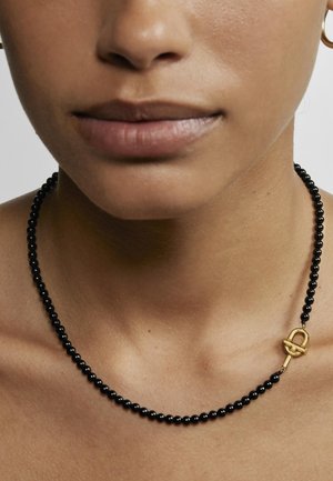 Collar de cuentas negras con una textura suave, que presenta un cierre de toggle dorado. Se lleva cerca de la clavícula sobre la piel desnuda.