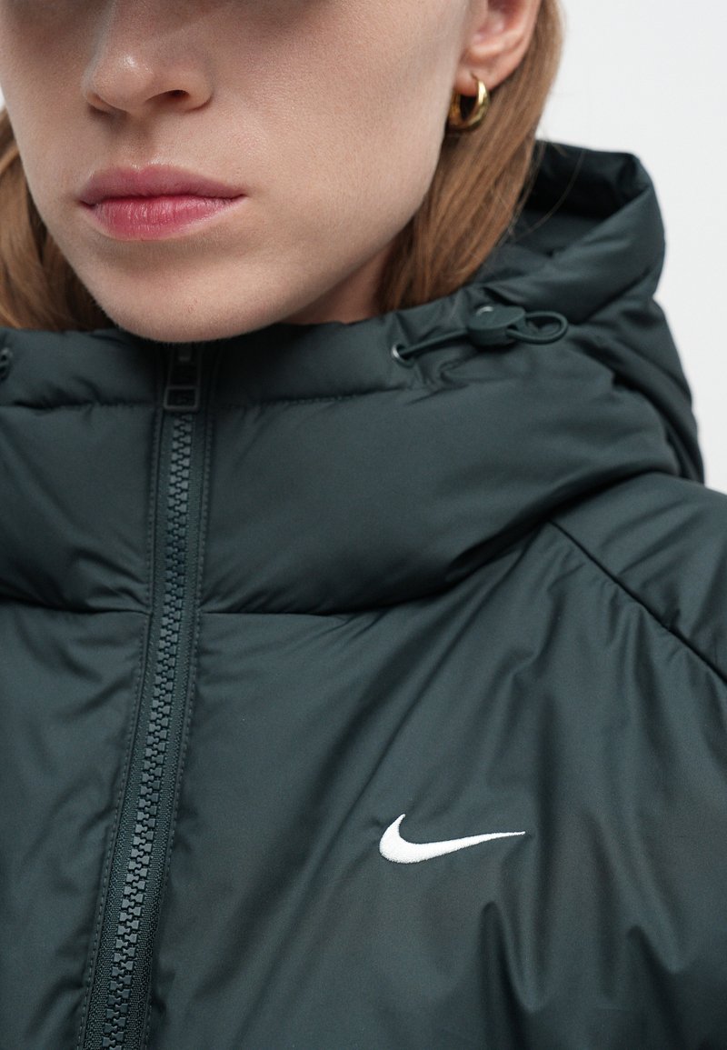 Dark green padded jacket with a high collar, front zipper, and a white Nike logo on the chest. Material appears smooth with a textured finish.