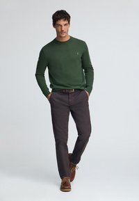 Grüner Strickpullover, braune Chinos und braune Lederhalbschuhe mit Schnürsenkeln. Der Pullover hat einen Rundhalsausschnitt und ein kleines Emblem auf der Brust.