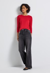 Frau steht an einer weißen Wand, trägt ein rotes Langarmoberteil, schwarze weit geschnittene Jeans und schwarze flache Schuhe und blickt zur Seite.