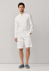 Hombre con camisa blanca de manga larga y cuello mandarín, pantalones cortos blancos hasta la rodilla y alpargatas azules, posando frente a un fondo gris.