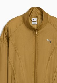 Giacca leggera color sabbia con colletto alto, chiusura con zip e dettagli di cuciture verticali. Include il logo argentato della Puma sul petto.