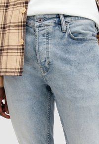 Jeans de mezclilla azul claro con un ligero desgaste, cierre de botón y cremallera, y diseño de cinco bolsillos, combinados con una camisa de cuadros.
