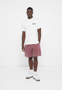 White cotton T-shirt with black logo, paired with pinkish-red corduroy shorts. White sneakers with gray and blue accents, plain background.