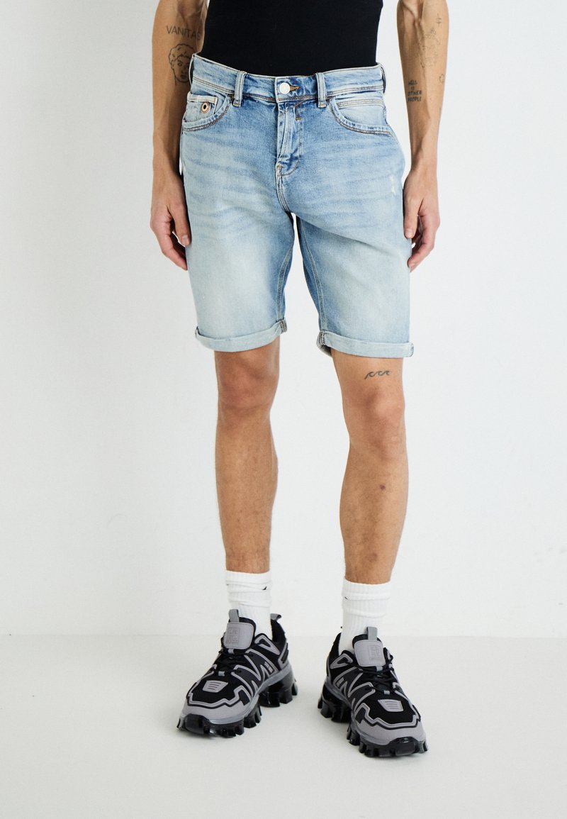 Shorts en denim de couleur bleu clair avec une coupe décontractée, des revers repliés et des coutures contrastées. Associés à des chaussures de sport noires et grises.