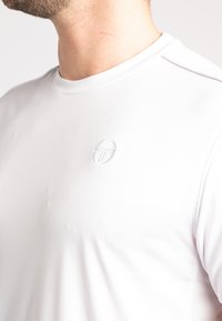 Vit idrottströja med rund halsringning och en subtil, texturerad logotyp på vänster sida. Tillverkad av fuktavledande tyg.