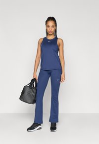 Marineblå treningstopp uten ermer og utsvingte bukser med Nike-logo, kombinert med svarte sneakers og en svart treningsbag. Glatt, elastisk stofftekstur.