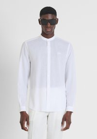 Camicia bianca di lino con colletto a stand, maniche lunghe e un logo discreto. Modello con occhiali da sole neri, abbinati a pantaloni chiari.