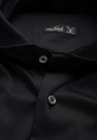 Schwarzfarbiges Hemd mit Stehkragen, das eine glatte Stoffstruktur aufweist, zwei Knöpfe an der Knopfleiste und ein Etikett mit einem gekreuzten Logo.