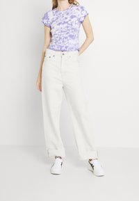 Camiseta de tie-dye morado con mangas cortas, jeans anchos de talle alto blancos y zapatillas blancas con detalles en negro, todo sobre un fondo liso.